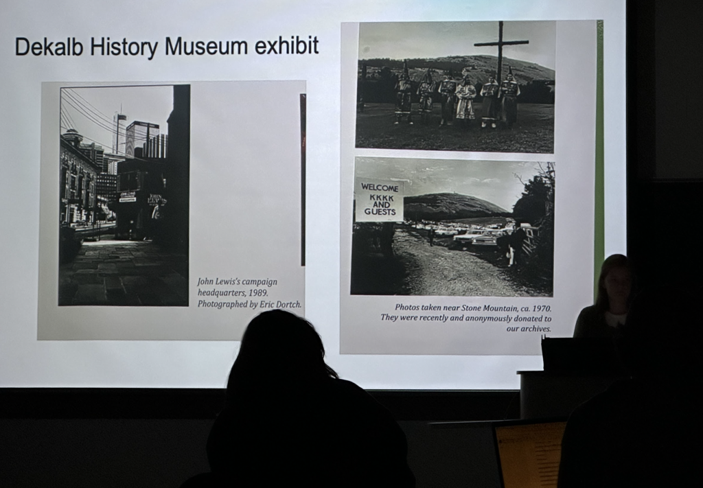 Student presents a slide to the class titled History Museum, A caption below a slide that displays three black and white images reads " photos taken near Stone Mountain, ca. 1970. They were recently and anobnymously donated to our archives." The images depict a sign on the road to Stone Mountain that reads "Welcome KKK and guests", another depicts 6 figures robed in KKK outfits that mask their identity and they are supporting a large wooden cross